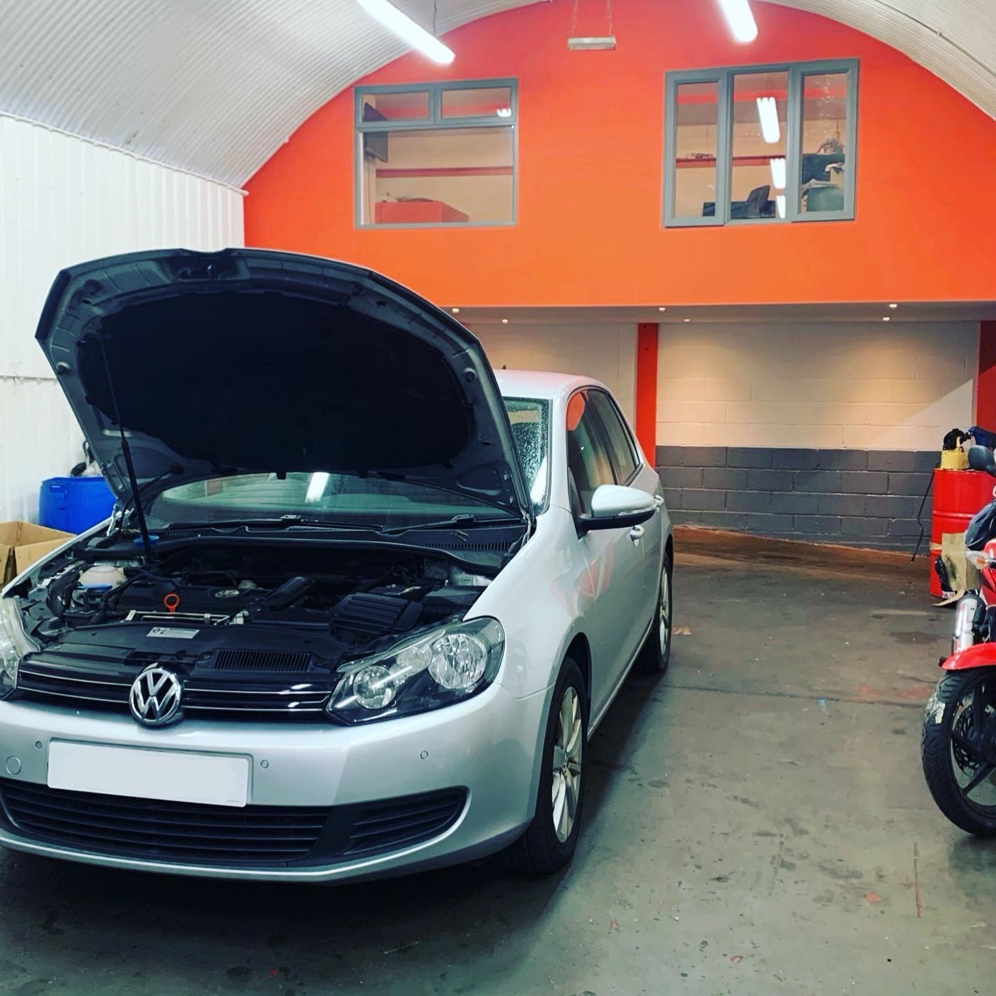 A silver Volkswagen Golf with its bonnet up in the Witan Motors workshop, next to a motorbike, beneath the orange arch wall.